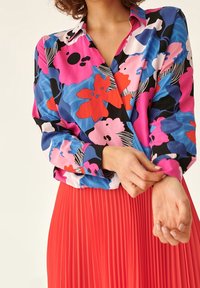 Donna che regola una blusa floreale a maniche lunghe con motivi rosa, blu, rossi e neri, abbinata a una gonna plissettata rossa a vita alta.