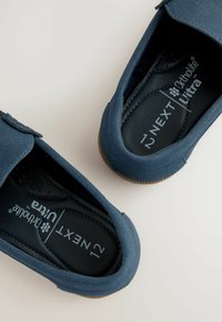 Des chaussures slip-on en suede bleu avec un bout arrondi et une semelle intérieure rembourrée noire, dotées d'une sangle à crochets et d'une semelle extérieure en caoutchouc marron.