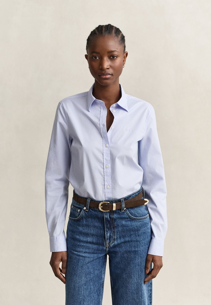 Femme aux cheveux tressés portant une chemise bleu clair boutonnée, rentrée dans un jean bleu avec une ceinture marron, debout devant un fond uni.