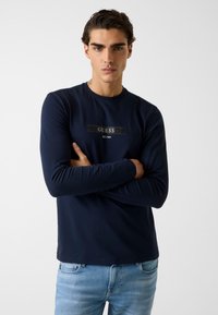 Granatowa koszulka z długim rękawem z czarnym prostokątnym logo z napisem "GUESS EST.1981" na piersi. Wykonana z mieszanki bawełny.