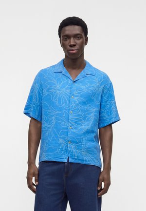 Bărbat stă în fața unui fundal simplu, purtând o cămașă albastră cu mâneci scurte și model floral și pantaloni bleumarin, privind înainte.