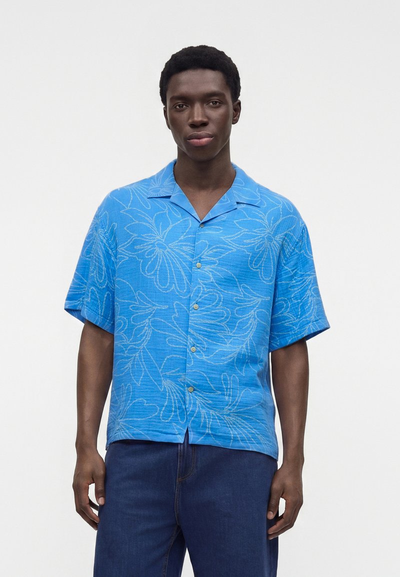 Homme debout devant un fond uni, portant une chemise bleue à manches courtes à motif floral et un pantalon bleu foncé, face à l'avant.