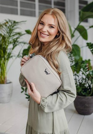 Béžová obdélníková taška se zipem, na jejímž přední strani se nachází černé logo. Model ji drží proti pozadí zelených rostlin.