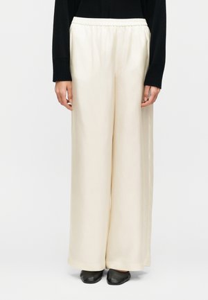 Άτομο που φοράει κρέμ παντελόνι με ψηλή μέση και φαρδιά ραφή, με ελαστική μέση, συνδυασμένο με μαύρο μακρυμάνικο τοπ και μαύρα επίπεδα παπούτσια σε λευκό φόντο.