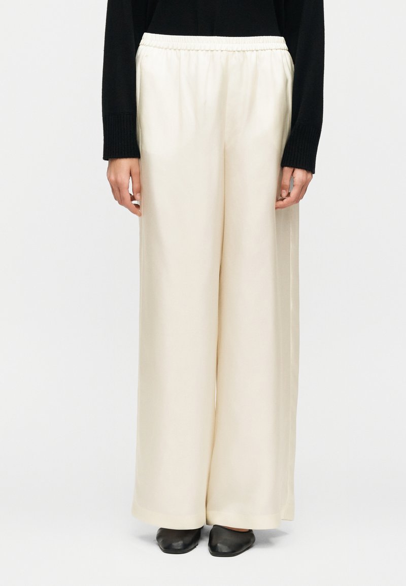 Personne portant un pantalon crème taille haute à jambes larges avec une ceinture élastique, associé à un haut noir à manches longues et des chaussures plates noires sur un fond blanc.