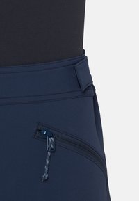 Close-up van de tailleband van marineblauwe broek met een schuin geplaatste ritszak en stoffen trekker aan de rits.