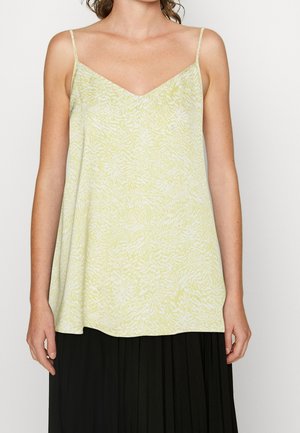 Woman wearing a light green patterned sleeveless V-neck top with thin straps and a black pleated skirt against a plain background.