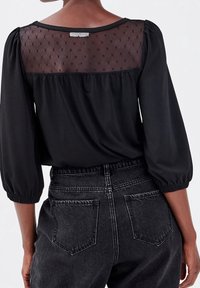 Blouse noire avec un panneau arrière en tulle à pois, manches trois-quarts volumineuses et coupe ample. Associée à un jean foncé taille haute.