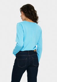 Vaaleansininen pitkähihaisempi paita, joka on valmistettu pehmeästä kankaasta ja jossa on pyöreä pääntie. Yhdistetty tummansinisiin farkkuihin, joissa on oranssia tikkausta ja takataskut.