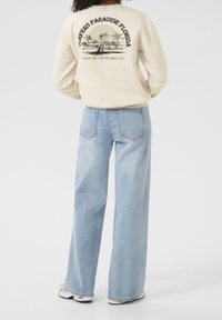 Sweatshirt beige clair avec imprimé graphique de palmiers et d'un voilier ; associé à un jean large bleu clair et des baskets blanches.