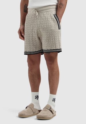 Gebreide beige korte broek met zwarte bies en trekkoort, gedragen met witte sokken met zwart monogram en beige slip-on schoenen met gesp.