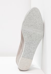Semelle d'une chaussure en daim beige avec une semelle extérieure en caoutchouc blanc texturé, inclinée contre un mur et un sol blancs.