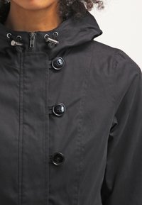 Czarna wodoodporna kurtka z przednim zamkiem błyskawicznym, kapturem i czterema dużymi czarnymi guzikami. Gładka tekstura z widocznymi detalami szwów.