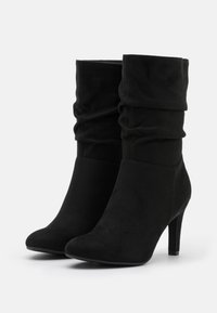 Botines negros de tobillo hechos de suave ante, con una parte superior arrugada, punta afilada y un tacón delgado. Textura lisa sin hardware visible.