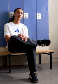 Valge T-särk sinise Adidas'i logoga, mustad teksad ja mustad tossud valgete triipudega, istub puidust pingil lukustatud kapide kõrval.