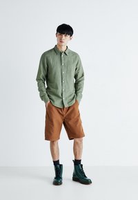 Camicia button-up verde in lino, pantaloni corti marroni e stivali verde con lacci. La camicia presenta un colletto a punta e cuciture visibili.