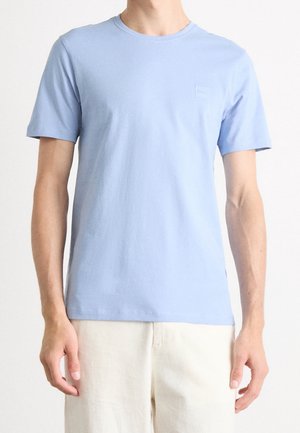 Mann trägt ein schlichtes hellblaues T-Shirt mit kurzen Ärmeln und hellbeige Hose, steht vor weißem Hintergrund.