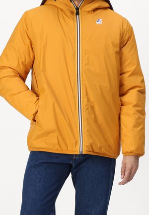 Personne portant une veste à capuche jaune avec fermeture éclair devant et un jean bleu, main gauche dans la poche, vue de la mi-cuisse au cou.