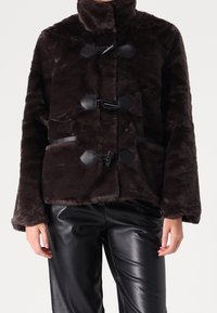 Brązowa kurtka z imitacji futra z wysokim kołnierzem, zapięciami na troki i teksturowaną powierzchnią; zestawiona z czarnymi skórzanymi spodniami.