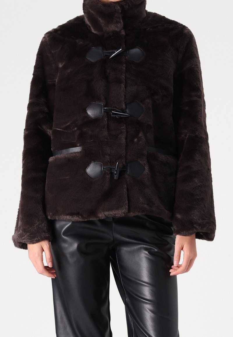 Brązowa kurtka z imitacji futra z wysokim kołnierzem, zapięciami na troki i teksturowaną powierzchnią; zestawiona z czarnymi skórzanymi spodniami.