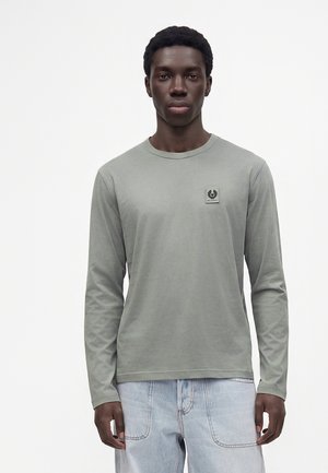 Junger Mann trägt ein hellgraues Langarmshirt mit kleinem Logo auf der Brust und hellblaue Jeans, steht vor einem einfarbigen Hintergrund.