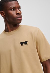 Bežist puuvillane T-särk, lühikeste varrukatega ja ümmarguse kaelusega, millel on mustade prillide graafika ja "KARL LAGERFELD" tekst tikandina.