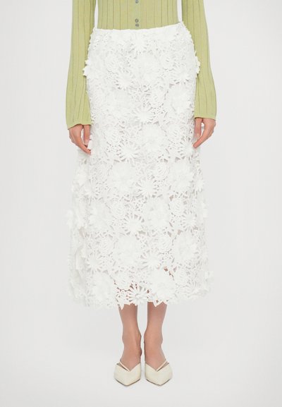 Witte kant midi rok met bloemen uitsnijdingen en gedetailleerde versieringen, gecombineerd met een groene ribbed blouse met lange mouwen en crèmekleurige spitse flats.