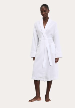 Femme debout pieds nus, portant un peignoir blanc texturé mi-long avec manches longues et ceinture nouée à la taille.
