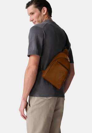 Brown leather sling bag with multiple zippered compartments, soft texture, and a streamlined design, worn over a grey sweater.
