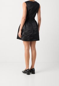 Abito in satin nero con corpetto aderente e gonna svasata, dotato di zip sul retro e dettagli plissettati. Indossato con scarpe nere con tacco a blocco.