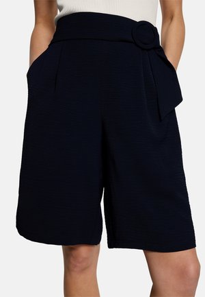 Shorts noirs taille haute arrivant au genou, dotés de poches latérales et d'une ceinture large avec une boucle ronde, portés avec un haut blanc sans manches.