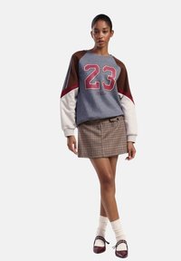 Felpa grigia con design rosso "23" e maniche marroni, abbinata a una gonna a quadri marroni. Calze bianche fino al ginocchio e scarpe marroni completano il look.