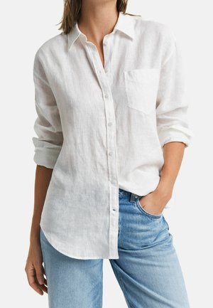 Woman wearing a white button-up long-sleeve shirt with rolled cuffs and light blue jeans, one hand in pocket, against a plain background.