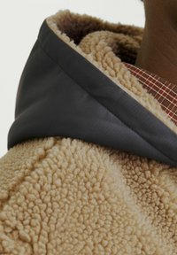 Beige fluffig tyg med en texturerad, mjuk yta. Huvud delen har en kontrasterande mörkgrå satin kant. Det inre lagret visar ett rutmönster.