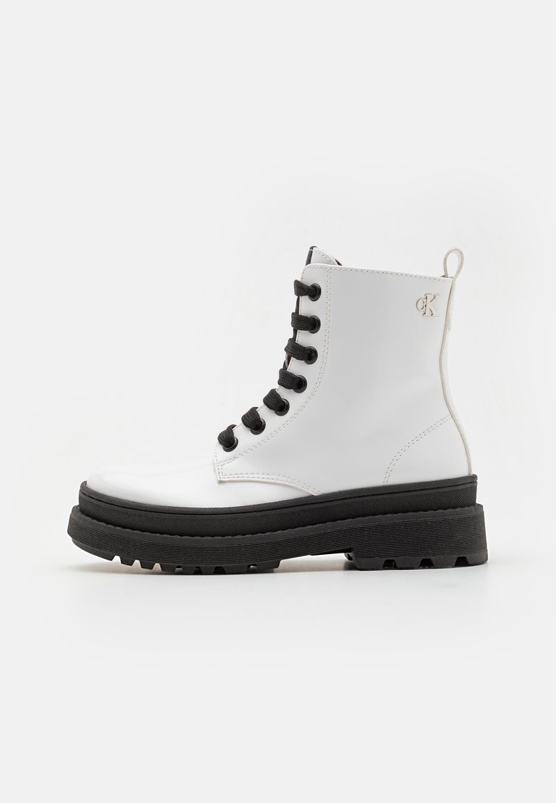 Botín de ante blanco con cordones negros, suela gruesa negra y un tacón pequeño. Presenta un acento de logo en la parte trasera.