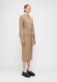 Abito in maglia marrone chiaro con texture a coste, maniche lunghe e vita legata. Abbinato a scarpe platform nere e spesse e calze beige.