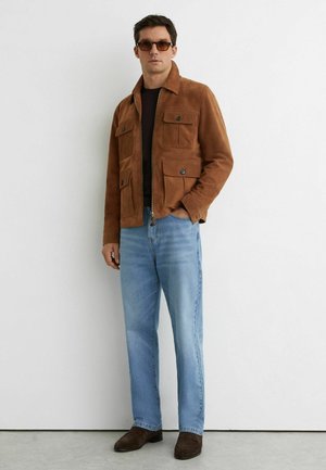 Muž nosiaci hnedú semišovú bundu s prednými vreckami, čiernu košeľu, svetlomodré džínsy, hnedé topánky a slnečné okuliare, stojaci pri bielom múre.