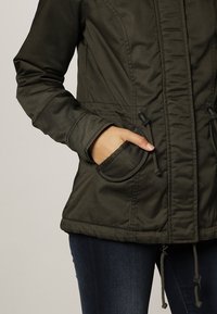 Osoba w oliwkowej kurtce z ręką w przedniej kieszeni, prezentująca sznurki kurtki oraz ciemnoniebieskie dżinsy.