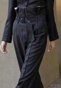 Svarta skräddarsydda byxor med ett brett bälte, med en slät yta och hög midja. Designen inkluderar knappdetaljer och en skarp silhuett.