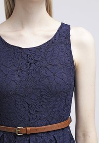 Marineblauwe kanten jurk met een bloemenpatroon, ronde halslijn en een bruine leren riem in de taille. Glad textuur en een aangesloten silhouet.