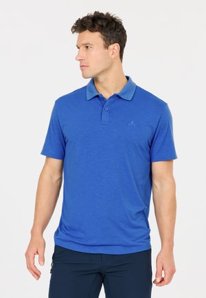 Mann trägt ein blaues Poloshirt mit kurzen Ärmeln und marineblaue Hose, blickt nach links vor einem schlichten weißen Hintergrund.