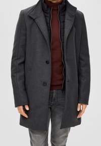 Cappotto in misto lana grigio con colletto a punte, due bottoni frontali e uno strato interno isolato nero con zip. Indossato sopra un maglione bordeaux.