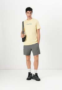 Camiseta de algodón amarilla con logo, pantalones cortos texturizados grises, zapatillas negras y calcetines blancos. El modelo sostiene una bolsa negra sobre un hombro.