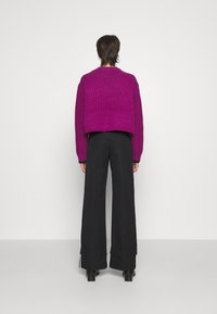 Persona de pie de espaldas, vistiendo un suéter corto morado, pantalones anchos negros con dobladillos enrollados y zapatos de tacón negros.