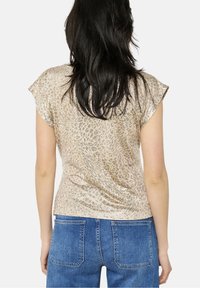 Krátký rukáv halenka v béžové barvě s jemným zvířecím potiskem, strukturovanou látkou a volnějším střihem na zádech, kombinovaná s modrými džínami.