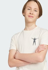 Camiseta de algodón color crema con un gráfico de una figura en silueta con los brazos extendidos en el lado izquierdo del pecho. Diseño de mangas cortas y cuello redondo.