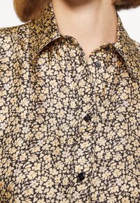 Chemise à boutons à motifs floraux, avec un fond marron foncé orné de petites fleurs jaunes et blanches, des boutons noirs et un col classique.