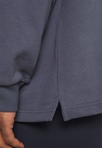 Tejido de sudadera gris oscuro con cuello y puños acanalados, que presenta aberturas laterales en el dobladillo para mayor comodidad y libertad de movimiento.