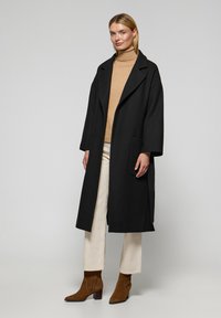 Abrigo de lana negro con un amplio cuello y dos bolsillos frontales, llevado sobre un jersey de cuello alto beige y pantalones color crema, complementado con botas marrones de tobillo.