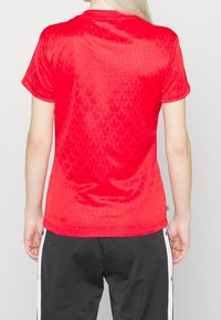 Červené sportovní tričko s krátkým rukávem, vyznačující se texturovaným vzorem tonálních listových tvarů, vyrobené z hladkého, lesklého materiálu.